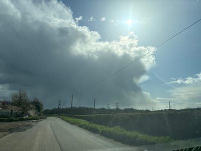 This image depicts a rural road in Smallfield, United Kingdom, under contrasting weather conditions. The sky is divided, with a bright sun visible through clear blue sky and scattered white clouds on the right, while a large, dense, dark grey storm cloud occupies the left and central upper portions of the frame. A single power line stretches diagonally across the upper-middle of the image.

The asphalt road, appearing damp, curves gently to the left in the foreground and midground. To the left of the road, a muddy or dirt area transitions to a metal fence. Behind the fence, a mound of earth or aggregate is visible, with a yellow excavator arm partially present on the far left. Deciduous trees, some bare and others bearing white blossoms, line this side. A small, illegible white sign is affixed to the fence.

To the right of the road, a neatly trimmed green hedge runs parallel to the asphalt. Beyond the hedge, a line of bare trees forms a distant treeline. Several large electricity pylons (transmission towers) are visible against the distant horizon and skyline, and at least two wooden utility poles are present closer to the road. No individuals or vehicles other than the presumed camera vehicle are present. The scene is illuminated by strong sunlight from the upper right, casting a bright glare.
