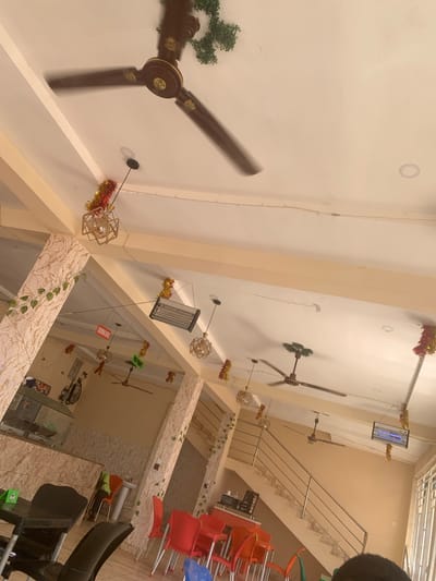 The image shows the interior of a building in Ungwan Liman, Nigeria. The space appears to be a commercial establishment, possibly a restaurant or cafe. The scene is captured from a high angle, looking down. There are multiple ceiling fans with brown blades, and some are decorated with green foliage or festive tinsel. The walls are light-colored. There are multiple stairs in the building. Tables and chairs are visible, with chairs stacked on some tables. There is a mix of seating colors, including red, blue, and green. There is a mirror with some decor on it.