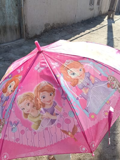 An open, pink umbrella with cartoon princess characters, specifically Sofia the First, is displayed outdoors in a city environment. The umbrella features images of Sofia and another princess, along with butterflies, flowers, and a bird. The background shows a dirt road and a building with a weathered facade. The scene is set in the city of Altinkul, Uzbekistan. The lighting suggests it is daytime, with shadows cast on the ground from a nearby pole.