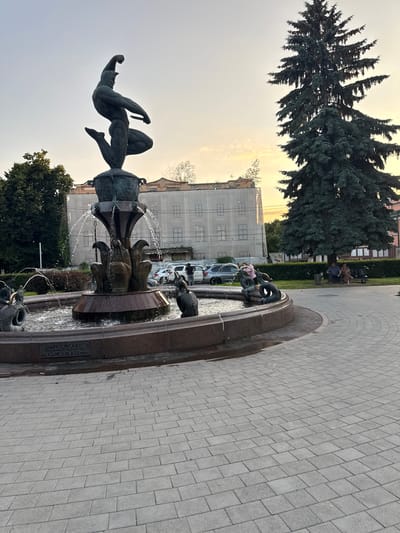 The image captures a fountain adorned with a dynamic sculpture of a man in mid-leap, possibly depicting an athlete or mythical figure. The fountain itself is multi-tiered with water cascading from several spouts, and features decorative elements resembling stylized plants and possibly animal heads at the base. In the background, a large evergreen tree stands prominently. Adjacent to the fountain, several people are visible; some are seated on a bench under the tree, while others are near parked cars. The environment includes a paved walkway and a building undergoing renovation, indicated by scaffolding and protective coverings. The sky suggests it is either sunrise or sunset, with soft, warm light illuminating the scene. A plaque on the fountain's base appears to contain text, likely identifying the artwork or its location. The overall scene conveys a public space, possibly a park or town square, in the city of Khamkhi, Russia.
