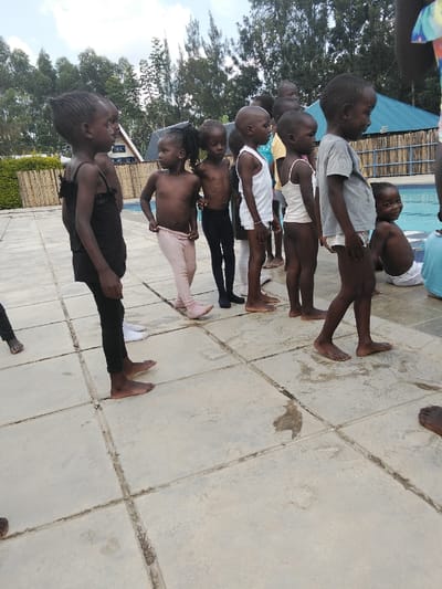 The image displays a scene in Malaha Isongo Makunga ward, Kenya, featuring approximately ten young children of African descent gathered on a light-colored, wet-splashed paved area adjacent to a swimming pool. Most children, estimated to be between four and eight years old, are standing in a loose, irregular line facing towards the right side of the frame.
From left to right, visible individuals include: a child in a black tank top and black leggings; a bare-torsoed child in light pink leggings, slightly pulled down; a bare-torsoed child in black leggings; a child in a white sleeveless undershirt and white briefs; and a child in a grey t-shirt and white briefs. All children on the paved surface are barefoot. One child is seated at the edge of the blue-tiled swimming pool on the far right, wearing white briefs and looking directly at the camera with a smile.
The immediate background behind the children features the blue-tiled swimming pool and a low, light-colored wooden or bamboo fence. Beyond the fence, numerous tall, green trees dominate the upper background. A building with a dark blue or green sloped roof is partially visible behind the trees on the right side. The sky is partially cloudy and bright.