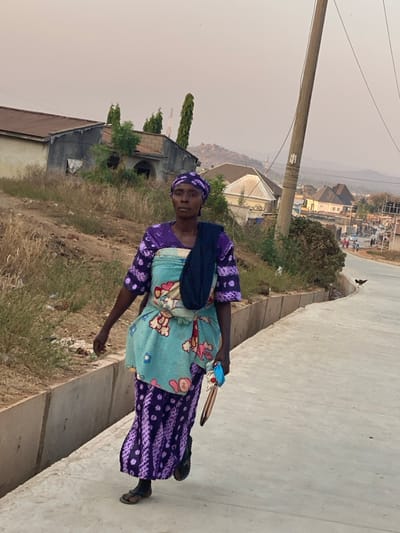 An elderly woman stands on a concrete road. She wears a patterned purple dress with a light blue fabric sling across her torso and a dark blue bag over her shoulder. The background shows a landscape with houses and a utility pole. The sky is hazy. She looks towards the camera. Her expression is serious. The environment appears rural or suburban.