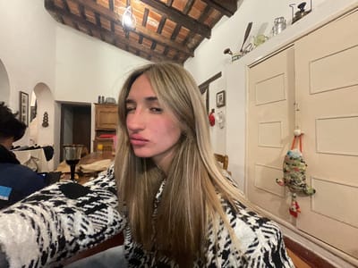 The image captures a selfie taken indoors, likely in a home environment in El Rodeo, Argentina. A young woman with long, blonde hair and a nose piercing is the primary subject, posing with her lips pursed. She is wearing a black and white patterned sweater. Her face is turned slightly to the left, with her eyes partially closed, suggesting she might be winking or attempting a playful expression.

The background reveals a room with a rustic aesthetic. A wooden ceiling with exposed beams and terracotta tiles is visible at the top. On the walls, there are framed pictures and decorative items, including a vintage oil lamp on a shelf above a cream-colored door. A stuffed animal toy in a camouflage pattern hangs from a hook on the door. To the left of the woman, a glimpse of another person is visible, partially obscured, wearing a dark jacket with a blue stripe. A table with a glass of red wine and some paper bags is also present in the background, suggesting a casual gathering or meal. The overall impression is one of a relaxed, indoor setting, potentially a living room or dining area.