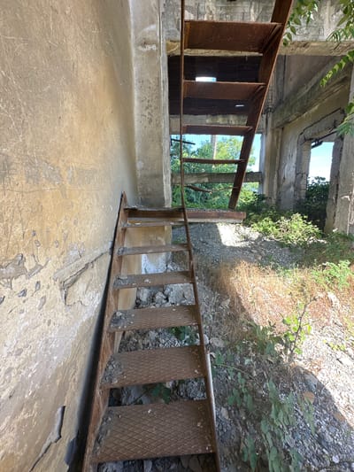 The image depicts an abandoned, partially collapsed concrete structure with a rusty metal staircase leading upwards. The walls of the structure are made of weathered concrete, with some sections showing peeling paint and graffiti. The ground is covered in rubble, dirt, and overgrown vegetation. Sunlight streams through openings in the roof and walls, illuminating the scene. The overall impression is one of decay and neglect. This is a view of an abandoned structure, possibly a former industrial building, located in or near the city of Rosen, Bulgaria. There are no people visible in the image.
