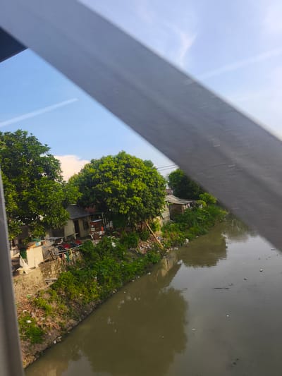 This image shows a view of a riverbank lined with informal settlements and dense vegetation. A wide, murky river flows through the scene, reflecting the sky and the surrounding foliage. On the riverbank, there are several small, makeshift houses constructed from various materials, indicating a low-income area. Litter and debris are visible along the bank and in the water, suggesting potential environmental concerns. Lush green trees, some of them large and mature, overhang the river and the settlements, providing shade and a natural element to the urban landscape. The sky above is partly cloudy with a bright blue expanse, suggesting it is daytime. The overall scene depicts a riverside community in a developing urban environment, likely in a tropical or subtropical region given the vegetation. The image is framed by a blurred, dark gray diagonal element, suggesting it was taken through a window or a similar barrier. The location in the city of Adidharma, Indonesia, would place this scene within a region known for its dense population and varying levels of infrastructure development, where informal settlements often develop along waterways.
