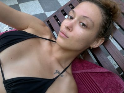 The image captures a woman relaxing on a dark wooden lounge chair, seen from an overhead, slightly tilted perspective. She is lying on her back, looking directly at the camera with a calm expression. Her head is positioned towards the lower left, and her torso, clad in a black bikini top, extends towards the upper right.
A dark pink or maroon towel is folded and placed beneath her head and upper back, resting on the wooden slats of the lounge chair. A small, stylized script tattoo, possibly reading "yoga," is visible on her left shoulder/collarbone area. She has brown hair, which appears to be pulled back, with a braid visible near her left ear. Her eyes are brown, and she has a natural skin tone with some visible freckles. A small golden hoop earring is visible in her left ear.
The background includes a distinctive checkered floor pattern of grey and white tiles, visible to the right and bottom of the frame. This, along with the lounge chair, suggests an outdoor or poolside setting, indicative of a leisure or vacation environment. The lighting is bright, consistent with daytime. The scene evokes a relaxed atmosphere, likely in Unawatuna, Sri Lanka.