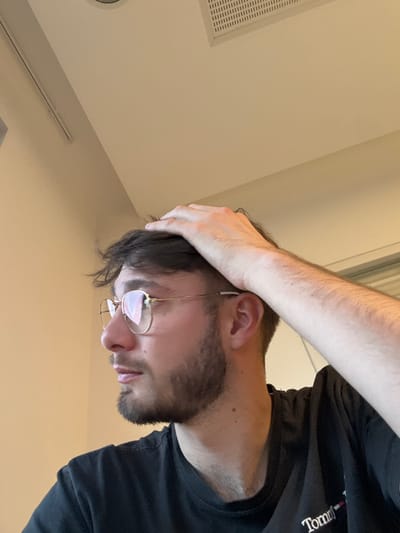 A young adult male is depicted in an indoor setting, viewed from his left profile, looking towards the left. His right hand is placed on the top of his head, with fingers resting in his dark brown, wavy hair. He has light skin, a trimmed beard, and wears gold-rimmed hexagonal eyeglasses with clear lenses; his lips are slightly parted. He is dressed in a black t-shirt featuring a partially visible white logo that includes the text "Tommy". The background comprises plain, light-colored walls and a matching ceiling. Visible on the ceiling are a square recessed vent with horizontal slats and a circular light fixture. This scene is located in the city of Lièpvre, France.