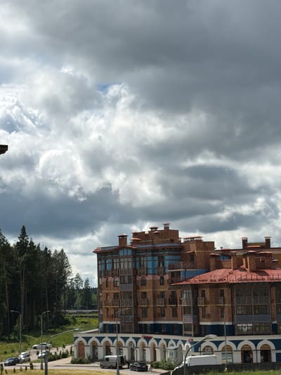 The image depicts a multi-story building with a red tiled roof and a brick facade, featuring decorative blue accents and numerous windows. The building appears to be a residential or hotel complex, possibly located in a suburban or semi-rural area. In front of the building, there is a road with several parked cars, including sedans and a van. A few streetlights are visible along the road. To the left of the building, a dense forest of tall pine trees forms a backdrop. The sky is overcast with heavy, dark gray clouds, suggesting a potentially stormy or overcast day. The architecture of the building, with its intricate brickwork and prominent chimneys, hints at a design that may be found in the Moscow Oblast region of Russia, specifically in a location like Николо-Урюпино, which is known for its suburban developments and surrounding natural landscapes. The overall impression is one of a modern building set against a natural, forested environment, under a dramatic sky.