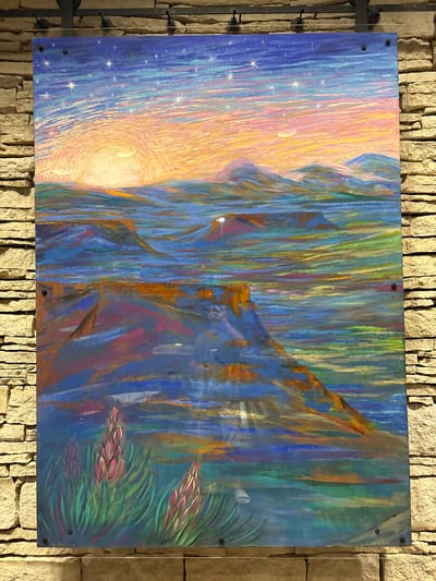 This is a photograph of a painting displayed on a stone wall. The painting is a colorful landscape with a sunrise or sunset scene. The sky is a gradient of deep blue at the top, transitioning through shades of pink, orange, and yellow towards the horizon, with numerous white starbursts scattered across it. A sliver of a crescent moon is visible in the upper right portion of the sky.
The middle ground of the painting depicts rolling hills or mountains rendered in blues, greens, and purples, with streaks of other colors suggesting light and shadow. There is a sense of depth created by the layering of these forms. In the foreground, a rugged, angled cliff face dominates the lower left quadrant, painted in rich blues and oranges.
Along the bottom edge of the painting, in the lower left corner, are three stylized pinkish-purple flower-like or seed-pod-like plants with green, spiky foliage. The overall impression of the painting is vibrant and expressive, with visible brushstrokes or textural marks.
The painting is mounted on a wall constructed of light-colored, rough-hewn stones. The wall has horizontal courses of these stones, creating a textured backdrop. The painting itself appears to be enclosed in a clear protective material, possibly acrylic or glass, and is attached to the wall with visible black screws or fasteners in the corners. Metal brackets are mounted at the top of the wall, supporting hanging wires that hold the painting. The scene is well-lit, with light sources casting a gentle illumination on the painting and wall. The location is identified as Unknown, United States of America.