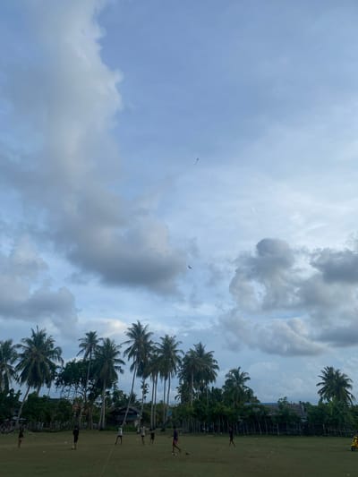 The image depicts an outdoor scene in Keudeublang, Indonesia, with a group of individuals playing soccer on a grassy field. Tall palm trees are prevalent throughout the landscape, lining the perimeter of the field and surrounding a few buildings in the background. The sky is partly cloudy, with large, dramatic clouds dominating the upper portion of the frame. Two kites are visible in the sky, one of which is a diamond-shaped kite. A bird is also flying in the sky. The overall atmosphere suggests a casual, leisurely activity taking place in a tropical environment.