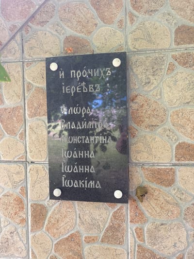 A black plaque with engraved text is affixed to a tiled wall. The text is in a Slavic script, likely Old Church Slavonic or a similar language. The plaque is mounted on a wall covered in irregularly shaped tiles in shades of beige, tan, and pinkish-brown, with grey grout. The lighting suggests it is daytime, with some dappled sunlight and shadows. A small, dried brown leaf is visible on the wall to the right of the plaque.
The text on the plaque appears to be a list of names or titles. The first line reads, "и прочиихъ". The second line reads, "іере́въ:". Subsequent lines appear to list names: "Флώра", "Владиміръ", "Константина", "Іωάνна", "Іωάνна", and "Іωакіма".
While the image itself does not provide direct information about the city of Puchły, Poland, similar plaques with such inscriptions are commonly found in Orthodox Christian churches or cemeteries. Puchły is known for its historical Orthodox church, the Church of the Transfiguration. Therefore, it is highly probable that this plaque is located at or near this church, serving as a memorial or record.