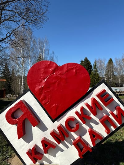 The image shows an outdoor sign in Galevo, Russia, featuring a large red heart and red Cyrillic letters on a white background. The sign appears to be located in a park or green space, as trees and grass are visible in the background. The sky is clear and blue.