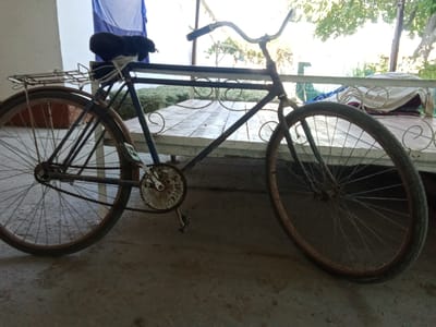 A rusty, old blue bicycle with a black seat and a rear rack is parked on a concrete floor. The bicycle has a chain and pedals. In the background, there is a wooden table with a white railing and some folded cloth items. The scene appears to be outdoors, possibly in a courtyard, with trees and foliage visible in the distance. The overall impression is one of disuse and aging. This scene is located in the city of Karagana, Uzbekistan.