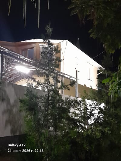 The image is a night-time photograph taken outdoors in Kara-Köl City, Kyrgyzstan. The scene features a multi-story building with a light-colored facade and several windows, partially obscured by trees and foliage. A covered walkway or balcony with ornate metalwork is visible in the foreground, illuminated by an overhead light fixture. The sky is dark, indicating nighttime. In the bottom left corner of the image, a watermark reads "Galaxy A12 21 июня 2026 г. 22:13", indicating the device used and the date and time the photo was taken.