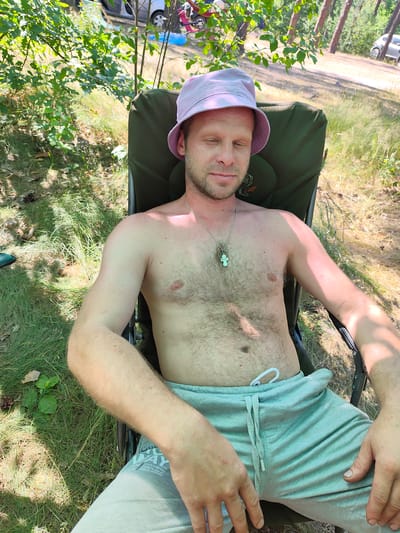 A shirtless man wearing a pink bucket hat and grey sweatpants sits in a green folding chair outdoors. He has his eyes closed and is leaning back in the chair. The man has light skin and is hairy on his chest and abdomen. He is wearing a silver chain with a pendant. The background shows trees, grass, and a dirt path with a car in the distance. The location is identified as Horods'ke, Ukraine.