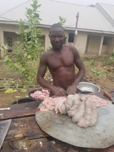 The image shows a man standing outside, wearing no shirt, in front of a wooden table. He is butchering animal intestines with his hands. He appears to be cleaning and preparing them for cooking or sale.  The intestines are laid out on a round wooden surface in front of him, along with other meat pieces in a black bowl. The man appears to be looking directly at the camera. In the background, two unfinished buildings are partially visible, along with a small green tree and some greenery in the surrounding area.  The scene is likely a residential or agricultural setting in Pati Ridge, Nigeria. The image suggests that the man is involved in traditional food preparation or trade.