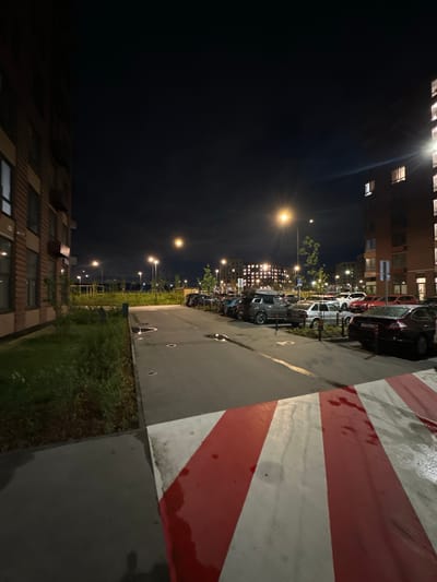 This is a night-time outdoor photograph taken in Laikovo, Russia. The scene depicts a parking lot adjacent to residential buildings. In the foreground, a wet asphalt surface is visible with red and white striped markings, possibly indicating a speed bump or a pedestrian crossing. To the left, a multi-story brick building with illuminated windows can be seen. A patch of green grass and vegetation borders the sidewalk. The middle ground features a street lined with parked cars. Various car models, including sedans and SUVs, are present. Streetlights illuminate the area, casting pools of light on the pavement and highlighting the wet surfaces from recent rain. In the background, more residential buildings with lit windows are visible, suggesting an urban or suburban environment. The sky is dark, typical of nighttime, with no visible celestial bodies.