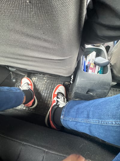 The image shows the interior of a vehicle, likely a car, from an overhead perspective. Two people are visible from the knees down. The person on the left is wearing gray pants, while the person on the right is wearing blue jeans. Both are wearing black socks and similar athletic shoes that are white with red and black details. The car interior is gray and black. A center console is between the passengers and contains cigarettes and a pen. The floor of the car is a black, patterned rubber mat. The location of Брусовeте, Bulgaria is not discernible in the image.