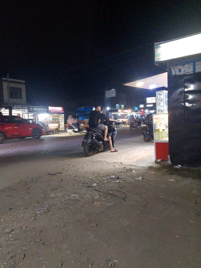 This is a nighttime, outdoor photograph taken in Gampongpaya, Indonesia. The scene depicts a street with several businesses and vehicles. In the foreground, a person is sitting on a motorcycle, facing away from the camera. The motorcycle is dark-colored and has a rider behind the driver.
To the left of the motorcycle, a red car is parked or driving by. Further back, there are multiple shops and stalls illuminated by bright lights. One stall has the text "WM BOUTIQUE" and another displays "WM DIMSUM". Another stall has the text "Nasi Goreng" and "Nasi Ayam". A bright white sign with the word "gopay" is visible above one of the businesses on the right side of the image.
The ground is unpaved and appears to be dirt with scattered debris. The lighting suggests it is nighttime, possibly late evening, with artificial lights from the shops and street illuminating the area. The weather appears to be clear. The overall atmosphere is that of a busy street in a commercial area at night.