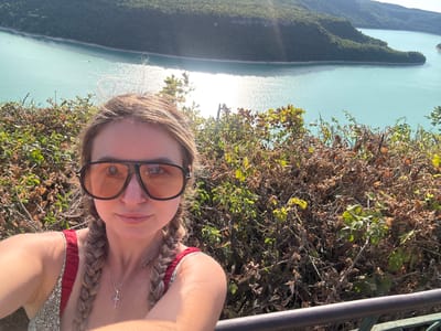 A young woman is taking a selfie outdoors with a body of water and forested hills in the background. The woman is wearing large, dark-rimmed sunglasses with orange lenses. Her blonde hair is styled in two braids. She is also wearing a red and white patterned top and a silver necklace with a cross pendant. The woman is positioned in the foreground, with a dense thicket of brown and green foliage to her right. Behind the foliage, a wide expanse of turquoise water is visible, reflecting the sunlight. The water is bordered by lush, green, tree-covered hills, characteristic of the region surrounding Moirans-en-Montagne, France. The sky above is clear and bright. A dark metal railing is visible along the bottom of the frame.