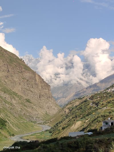 An aerial drone shot captures a scenic valley with a winding river in Yamling, India. The sky is blue with scattered clouds. A steep, rocky mountainside dominates the left side of the frame, while the right side features a terraced hillside dotted with small houses and lush greenery. A road snakes its way up the hillside. The river flows through the valley floor, bordered by green fields.