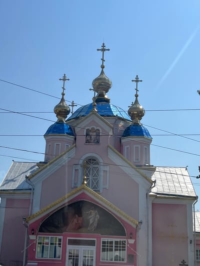 This image shows the exterior of a pink Orthodox church in the city of Kovel, Ukraine. The church features multiple golden domes topped with crosses, and blue tiled roofs. A large fresco is visible above the entrance. The sky is clear and blue, with power lines running across the frame. The building appears to be made of stucco and has white trim. The architectural style suggests it is a religious building.