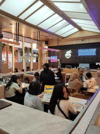 The image shows the interior of a sushi restaurant, likely in Kudus, Indonesia, as indicated by the "SUSHIKUN" sign. Several people are seated at tables, some are dining, while others are waiting to be served. A conveyor belt system is visible, typical for sushi restaurants, carrying plates of food. The decor features Japanese-inspired elements, such as hanging plants and signage with Japanese characters. The lighting is bright, with a modern, diffused ceiling light system. The overall scene depicts a lively dining environment.