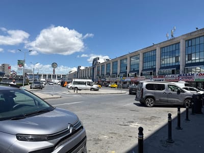 This image depicts a street scene in Bayrampaşa, Turkey. The sky is partly cloudy with a large white cloud towards the left. In the foreground is a gray Citroën car. The central focus is a traffic circle with various vehicles including cars, vans, and a yellow taxi. Pedestrian crossing signs are visible. A long, multi-story building with numerous windows and signage stretches across the background. Some signage visible includes "KONTUR KESAN" and "ISTANBUL." There are lamp posts and black bollards along the side of the road. People are visible in the background, but are not a prominent feature.