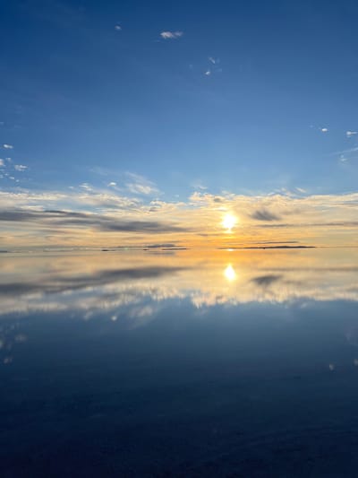 This image presents a panoramic view of the Salar de Uyuni within the Municipio Colcha K, Bolivia, at either sunrise or sunset. The scene is horizontally bisected by a sharply defined horizon line. The upper half displays a clear blue sky that gradually transitions to vibrant yellow and orange hues closer to the horizon. A brilliant sun, a central point of light, is positioned slightly to the right of the vertical midline. Various scattered clouds, ranging from wispy white to more diffused grey formations, are visible across the sky, particularly concentrated in the middle and lower sections of the sky. The lower half of the image is a highly reflective, mirror-like surface, consistent with a thin layer of water covering the salt flat. This surface provides a near-perfect reflection of the entire sky above, including the distinct sun directly below its actual position and the corresponding cloud formations. The immediate foreground of the reflective surface shows subtle textural variations, possibly indicating the underlying salt crust beneath the water. There are no individuals, animals, or man-made objects present in the frame. The scene's interaction is primarily optical, with sunlight and atmospheric elements reflected upon the vast, still surface.