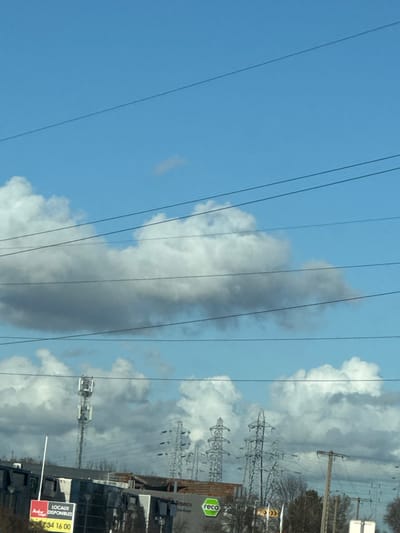 The image depicts an outdoor scene in Souffelweyersheim, France, under a clear blue sky interspersed with numerous white and grey cumulus clouds. The upper two-thirds of the frame are dominated by the sky and several dark, taut electrical wires running horizontally across the view.
In the lower portion of the image, an urban or industrial landscape is visible. On the left, a tall, grey cellular communication tower with multiple antenna arrays extends upwards into the clouds. Below it, a white commercial sign with red lettering reads "LOCAUX DISPONIBLES" (Available Premises) and displays the phone number "03 88 34 16 00." Adjacent to this sign are sections of dark-colored commercial or industrial buildings with flat roofs. One building prominently features a green circular logo with the word "reca" in white lettering.
Towards the center and right mid-ground, a series of large, grey lattice-steel high-voltage electricity pylons stand against the sky and clouds, supporting numerous power lines. Further right, a single dark wooden utility pole with crossarms and wires is present. The ground level below these structures shows sparse, leafless deciduous trees and shrubs, suggesting a cooler season. No individuals are visible, and no discernible actions or interactions are occurring within the frame. The perspective suggests the image was captured from a moving vehicle.
