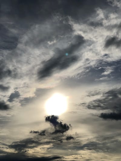 A cloudy sky is depicted, with a bright sun partially obscured by dark, wispy clouds. Some clouds exhibit iridescent colors, suggesting a halo effect around the sun. The image appears to be a photograph taken from the ground, looking upwards at the sky. There are no visible people, animals, buildings, or any other discernible objects or actions. The scene is natural and atmospheric, without any specific urban features identifiable. The location of Uriupino, Russia, is an unsupported assertion.
