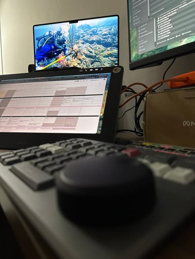 This media file shows a desk setup with multiple computer monitors, a keyboard, and various cables. One monitor displays a scuba diver with a lionfish in an underwater scene, likely a wallpaper. Another monitor shows a spreadsheet or calendar grid with text, some of which appears to be in Spanish. A portion of a keyboard with a large, dark knob and colored keys is visible on the right side of the image. Cables, including an orange one, are bundled together, and a small beige box with an infinity symbol is also present. The scene appears to be an indoor workspace, possibly a home office or studio. The overall impression is one of a functional, technology-filled environment. Visible text includes file names and application names like "DaVinci Resolve Studio," "iCloud Drive," and "Macintosh HD." The location context of Santa Marta, Colombia, does not provide specific visual cues within the image itself. There are no clear indications of time of day, weather, or overt emotions conveyed.