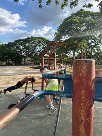 The image shows three men exercising on outdoor gym equipment in Parroquia Ocumare, Venezuela. In the foreground, a rusty metal structure with blue and red paint is visible. A shirtless man in black pants is using the equipment for a chest exercise. Behind him, another man in a white shirt and green shorts is stretching on the same structure. Further in the background, a third man is exercising on a different piece of equipment. The gym area is outdoors, with trees and a cloudy sky in the background. A painted wall and other gym equipment are also visible in the distance. The ground appears to be concrete.