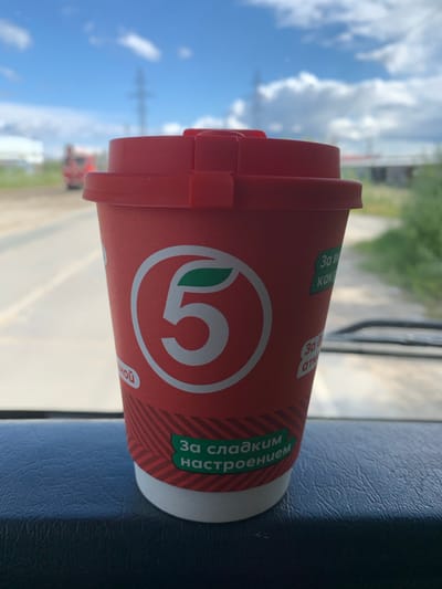 A close-up, eye-level shot shows a red disposable coffee cup with the logo of "Pyaterochka" (a Russian grocery store chain), which is a white circle with a green leaf and the number "5". The cup is placed on a dark blue textured surface, possibly the dashboard of a vehicle. The lid of the cup is also red. In the background, slightly out of focus, is a road, green vegetation, and a cloudy blue sky. A power line tower is visible in the distance. The context suggests this photograph was taken in Gubkinsky, Russia, given the presence of a Pyaterochka grocery store logo and the general environment.