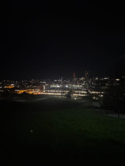 The image depicts a nighttime cityscape, likely taken from an elevated position on a grassy area. The city lights are visible in the distance, with the buildings appearing to be clustered together. The foreground consists of dark grass and possibly some wooden structures. The lights of the city are reflected off a street, and what appears to be a parkade with lighting. The angle and composition of the image suggest that it may have been taken from a hill or raised area overlooking the city. Given the overall look of the buildings, lights and green areas, it is most likely Sheffield, United Kingdom.