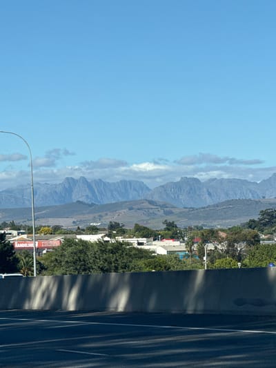 This media file captures a panoramic outdoor scene, likely taken from a vehicle or roadside viewpoint, looking towards a prominent mountain range. The image, oriented vertically, displays a clear blue sky with some scattered white clouds, particularly around the mountain peaks, indicating good weather conditions during the day.
In the mid-distance, a long, rugged chain of greyish-blue mountains stretches across the horizon, partially veiled by light clouds. The mountains feature distinct ridges and valleys. Below the mountains, the landscape transitions into a mix of green fields, trees, and scattered buildings.
In the foreground and mid-ground on the right side of the frame, there is a dark asphalt road or highway with clear white lane markings. Long, diagonal shadows are cast across the road surface and an adjacent barrier or wall, suggesting bright sunlight and possibly morning or late afternoon.
On the left side of the road, a commercial or residential area is visible, featuring green trees and buildings. Several red signs with white text are discernable. One sign clearly reads "Prestige Auctioneers" with "HANDELSHU" (likely part of "Handelshuis," meaning trading house) partially visible below it. Another red sign further back appears to have the text "TIENERS" or similar. A curved street light arm extends from the bottom of the frame towards the center.
The scene depicts a blend of urban infrastructure and natural landscape, consistent with areas near Bellville, South Africa, which offers views of surrounding mountains. No specific human activity or identifiable individuals are visible. The atmosphere is calm and clear.