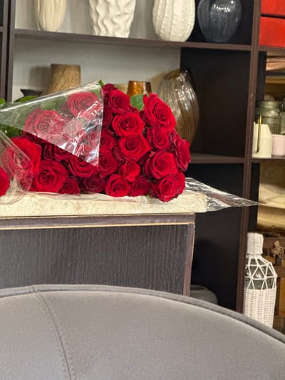 A bouquet of red roses is displayed on a dark wooden shelf. The roses are wrapped in clear plastic, and some of their petals are visible through the wrapping. The shelf is filled with various decorative objects, including glass vases and white textured items. The background shows more shelves with more items, suggesting a retail or display setting. A portion of a gray upholstered chair is visible in the right foreground, partially obscuring the view of the roses. The scene is brightly lit, with no clear indication of the time of day or weather. No text is visible in the image.