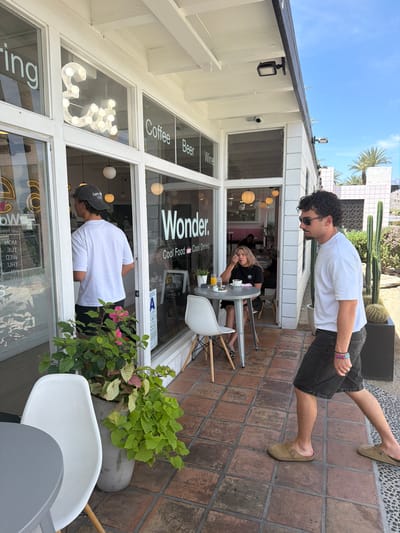 Here is a detailed description of the image:
**Scene:** The image captures the front exterior of a restaurant named "Wonder" during daylight hours, most likely located in Palm Desert, California, United States, judging by the desert flora and architectural style. The restaurant has a white facade with prominent window signage indicating "Coffee," "Beer," "Wine," and "Wonder. Cool Food Cool Drinks."
**People:** There are at least three people visible in the image:
1. A man, wearing a white t-shirt and dark shorts, walks outside the restaurant on a brick-paved area.
2. Another person inside the restaurant wearing a white T-shirt and baseball cap is visible at the window.
3. A woman is sitting at a table with a drink inside the restaurant.
**Objects:** Key objects in the scene include:
* The restaurant building with its signage and lighting.
* Chairs and tables, both inside and outside the restaurant.
* Plant pots with various plants near the entrance.
* The brick-paved ground.
* Other buildings and cacti in the background.
**Actions:**
* The man is walking past the front of the restaurant.
* Another man might be looking into the restaurant.
* The woman is seated at a table inside the restaurant.
**Environment:**
* The scene takes place outdoors on a sunny day.
* The background includes other buildings, palm trees, and cacti, indicating a desert environment likely located in Palm Desert, California.
