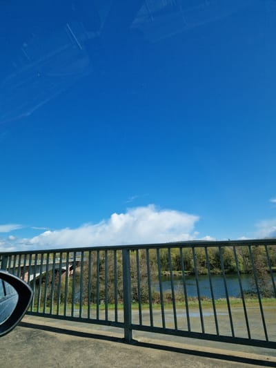 The image shows a view from a bridge in Obernburg am Main, Germany. The sky is bright blue with scattered white clouds. A grey metal railing runs along the right side of the image. Through the railing, a body of water and trees are visible. A car's side mirror is visible in the lower right corner. The surface to the right of the railing appears to be concrete.