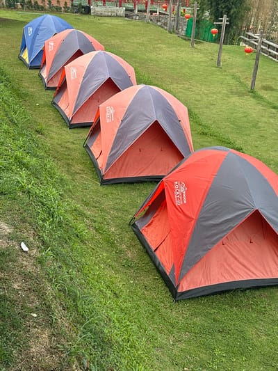 Six dome-style camping tents are pitched in a diagonal line on a green, grassy slope in Ban Mai Saha Sampan, Thailand. The leftmost tent is blue with yellow accents. The subsequent five tents are orange and dark gray, with "OUTDOOR ROCKY-II" branding visible on several. No individuals are present. The tents are arranged on a well-maintained lawn that slopes upward towards the background. In the background, beyond the tents, there are structures with red roofs and railings, resembling traditional architecture. Multiple red spherical lanterns hang from wooden posts and structures in the mid-ground and background. Additional green foliage and wooden fencing are visible on the right side of the frame. The ground immediately adjacent to the tents on the left exhibits wilder grass and some exposed soil.