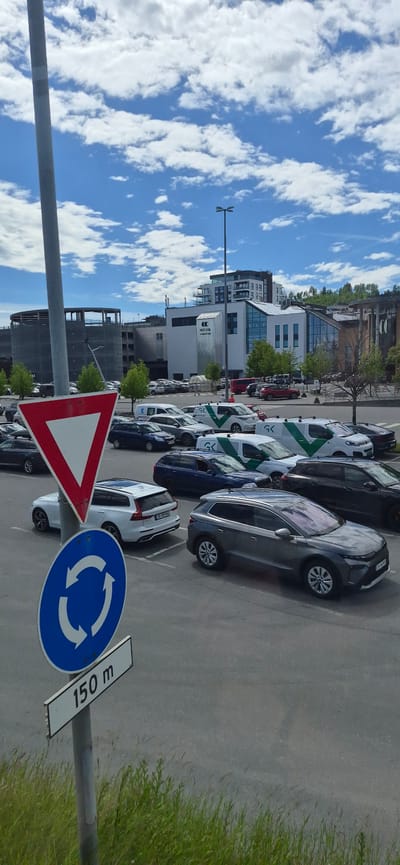 This is a vertical, full-color photograph taken on a sunny day with scattered clouds. The scene is a parking lot with numerous vehicles, including cars and vans. In the foreground, a traffic sign with a red triangle and a white circle with a red border indicates a "Give Way" or "Yield" sign. Below it, another blue circular sign with white arrows indicates a roundabout. A rectangular white sign beneath that reads "150 m".

Several cars are parked in the lot, including a white Volvo station wagon in the mid-ground, and a dark grey SUV in the foreground. Several white vans with green "GK" branding are also visible. In the background, there are modern buildings, including a multi-story white and glass structure with a prominent "10-71 (18)" sign. Some of these buildings appear to be under construction or renovation. The parking lot is paved with asphalt, and there is a strip of green grass and weeds along the bottom edge of the image. The sky is blue with puffy white clouds.

Based on the license plate of the white Volvo, "TD 89398", and the distinctive architecture of the buildings in the background, this image was likely taken in the city of Gjøvik, Norway, possibly near the Gjøvik Olympic Cavern Hall or the surrounding commercial area.