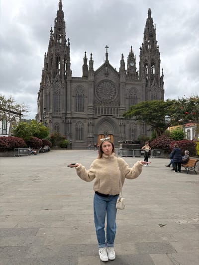 A young Caucasian female stands centrally in a large, paved public square, identified as the plaza in front of the Church of San Juan Bautista in Arucas, Gran Canaria, Spain. She has light blonde hair with sunglasses positioned on her head and wears a beige fleece zip-up jacket over a dark top, light blue cuffed jeans, and white platform-soled sneakers. A small cream-colored shoulder bag with a chain strap hangs from her left shoulder, and she holds a dark smartphone in her left hand. Her arms are extended outwards and slightly upward, palms open and facing up, with her shoulders shrugged and lips pursed, conveying a gesture of uncertainty.
Dominating the background is the Neo-Gothic Church of San Juan Bautista, a grand structure constructed from dark volcanic stone. It features two tall, intricately detailed spires, a large central rose window above the main entrance, numerous arched windows, and a visible clock on the right spire. The plaza itself is paved with grey flagstones.
To the left, beyond some low red bushes and metal barriers, multiple individuals are seated. To the right of the central subject, a woman stands with hands on her hips, facing towards the right. Further to the right, two older individuals are seated on a wooden park bench. Green trees and additional red-leafed bushes are visible around the plaza's perimeter. Stacked metal components or stage equipment are present on the right side of the plaza near the church. The sky above is completely overcast with dense grey clouds.