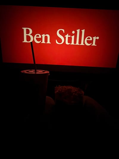 The image shows a museum exhibit. On the left, a vertical red sign displays the name "Ben Stiller" in white, sans-serif font. A thin black support extends from the sign towards the right, where a spotlight illuminates a bust of a person. The bust is placed in a dark environment, obscuring details of the surrounding area. This photograph was taken inside the Museum Pfalzgalerie Kaiserslautern, in Kaiserslautern, Germany.