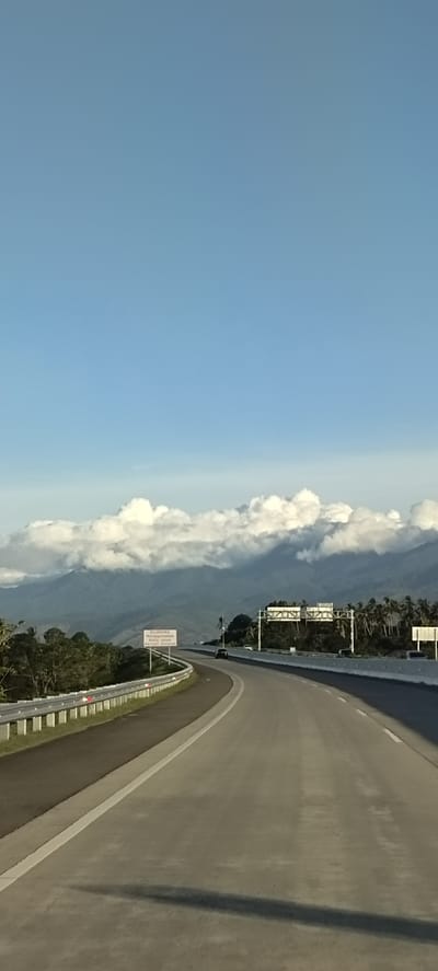 The image shows a highway curving through a mountainous landscape under a clear blue sky with scattered white clouds. In the distance, large, verdant mountains are partially obscured by a layer of fluffy white clouds. A sign in Indonesian, "DILARANG Menggunakan Kertsu Bararat," is visible on the left side of the road. The highway has a concrete surface with white lane markings and a metal guardrail. Palm trees and other lush vegetation line the sides of the road in certain areas. A few vehicles are present on the highway. The overall scene suggests a scenic route likely within or near Baksukon, Indonesia, given the presence of Indonesian language signage and the tropical vegetation.