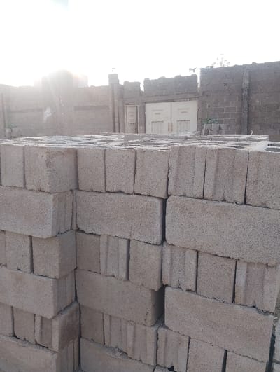 Stacks of gray concrete blocks fill the foreground. Each block has two vertical rectangular holes. Behind the blocks, a weathered brick or cinder block wall surrounds a yard. A closed, double gate is visible within the wall, flanked by single doors. The sky is partly visible in the upper portion of the image, with a bright light source partially obscured by the wall, casting a hazy glow.