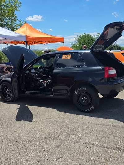 An outdoor scene, possibly at a race track or car show, features a black, modified hatchback car with its hood and trunk open, parked on asphalt. The car appears to be a drag racing vehicle, evident by the roll cage visible inside, racing tires with "Hoosier" branding, and a stripped-down interior. The car's driver-side door is also open, revealing more of the interior. To the left of the car, a white canopy is partially visible, and next to it, an orange canopy is prominently displayed. In the background, a person in a white cap and another person are visible near the canopies. Trees and a blue sky with scattered clouds form the backdrop. The car is positioned on a cracked asphalt surface, with a shadow cast from the car. The scene is set in Akatsievo, Bulgaria.