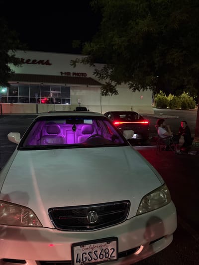 The image shows a white Acura sedan parked in front of a Walgreens store at night in Antelope, California. The car's interior is illuminated with purple ambient lighting. Two people are sitting in chairs outside the store, to the right of the car. A black car is parked behind the Acura. The Walgreens store has a red awning and a sign that says "1-HR PHOTO". Trees are visible in the foreground and background.