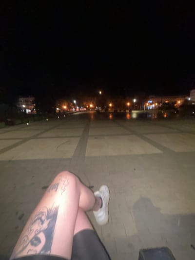 The image depicts a low-angle, night-time perspective from a seated position, focusing on the photographer's tattooed left leg and white athletic shoe. The leg exhibits a prominent tattoo of a creature with sharp teeth, possibly a dragon or monster, extending down the thigh. The background reveals a city square or promenade in Siedlce, Poland, characterized by patterned paving stones, streetlights casting warm glows, and illuminated buildings in the distance. The ground appears slightly wet, reflecting the lights, suggesting recent rain or damp conditions. The overall scene is dark, with artificial lighting creating a moody atmosphere, and the focus is on the personal element of the tattoo and the broader urban environment.
