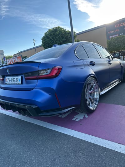 A blue BMW with chrome wheels is parked in a purple parking spot marked with white handprints. The license plate number is ZH-89289. The car is located near a building with "Coop City", "DENNER", and "C&A" signage. The image was likely taken in Volketswil, Switzerland.