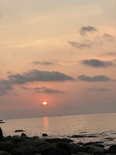 A wide shot depicts a sunset scene over the ocean in Preah Sihanouk, Cambodia. The sky, occupying approximately the upper two-thirds of the frame, displays a gradient of soft orange, pink, and pale yellow hues, interspersed with scattered dark grey and light grey stratocumulus clouds. The sun, a prominent semi-circle of intense yellow and orange light, is positioned low on the horizon in the left-center of the image, partially obscured by a cloud.

The calm sea surface, visible in the lower half of the frame, reflects the sun's light in a vertical, shimmering orange-yellow path extending from the horizon towards the viewer. A small, dark-hulled vessel is faintly visible on the distant horizon line to the right of the sun's reflection. The immediate foreground, at the bottom of the frame, consists of a rocky shoreline composed of numerous dark, irregularly shaped boulders, some appearing to be covered in patches of green algae. No individuals or discernible actions are present in the scene.